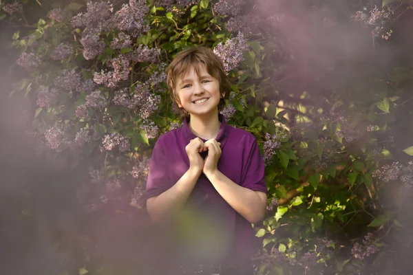 Ganska snäll pojke ler i lila träd och visar kärlek hjärtsymbol av handflatorna i sommarträdgården — Stockfoto