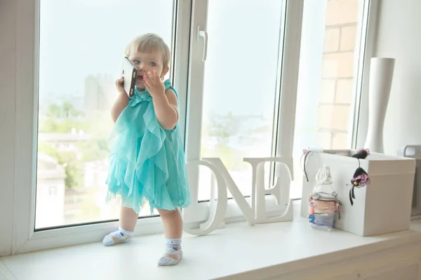 Meisje van de baby van een jaar praten via de telefoon in de buurt van het venster — Stockfoto