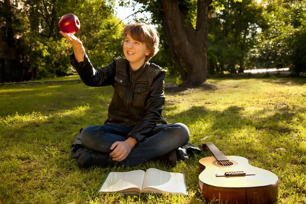 Adolescente ao ar livre com uma guitarra, um livro e uma maçã no pôr do sol no parque se divertindo — Fotografia de Stock