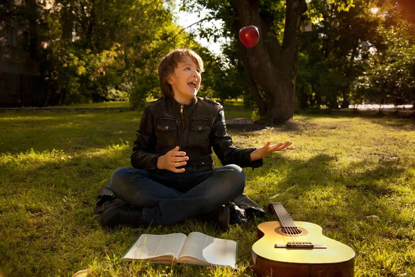 Adolescente ao ar livre com uma guitarra, um livro e uma maçã no pôr do sol no parque se divertindo — Fotografia de Stock