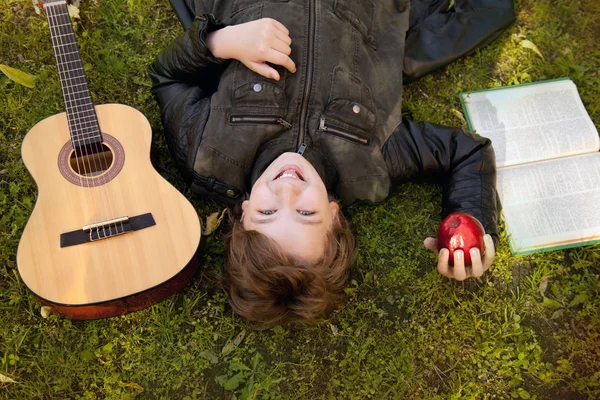 Adolescent garçon à l'extérieur avec une guitare, un livre et une pomme sur le coucher du soleil dans le parc avoir du plaisir — Photo