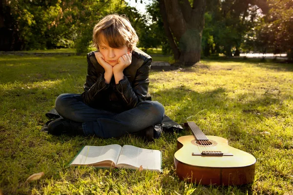 Tiener jongen buiten met een gitaar, een boek en een appel op zonsondergang in het park plezier — Stockfoto