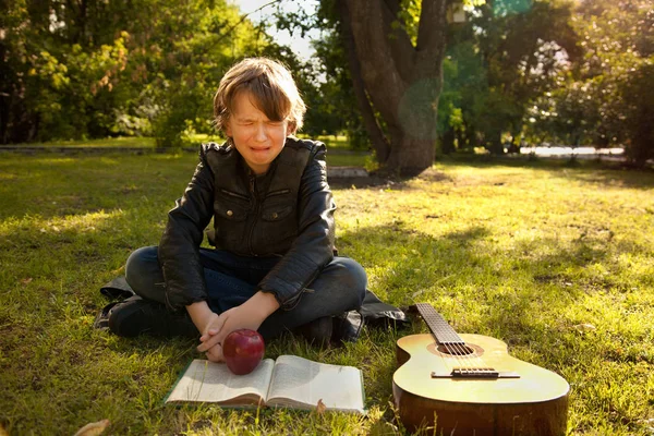Adolescente ao ar livre com uma guitarra, um livro e uma maçã no pôr do sol no parque chorar — Fotografia de Stock
