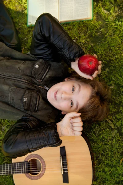 Adolescente ao ar livre com uma guitarra, um livro e uma maçã no pôr do sol no parque se divertindo — Fotografia de Stock