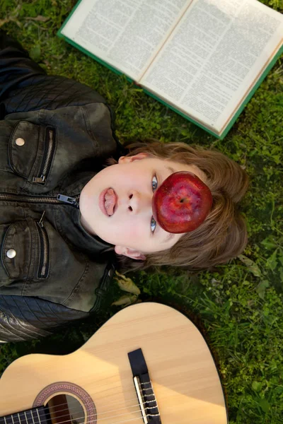 Adolescente ao ar livre com uma guitarra, um livro e uma maçã no pôr do sol no parque se divertindo — Fotografia de Stock