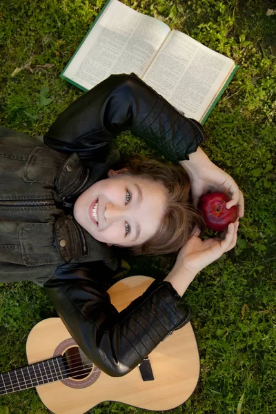 Teen boy outdoors with a guitar, a book and an apple on sunset in the park having fun — Stock Photo, Image