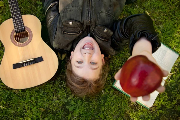 Teen chłopiec na zewnątrz z gitarą, książki i jabłko na zachód słońca w parku zabawy — Zdjęcie stockowe