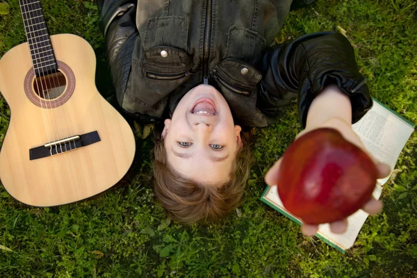 Adolescente ao ar livre com uma guitarra, um livro e uma maçã no pôr do sol no parque se divertindo — Fotografia de Stock