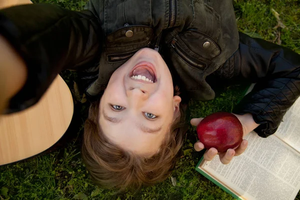 Adolescente al aire libre con una guitarra, un libro y una manzana al atardecer en el parque divirtiéndose y haciendo selfie — Foto de Stock