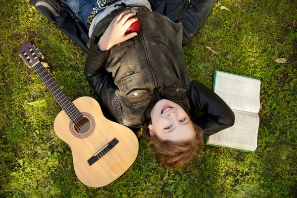 Мальчик-подросток на улице с гитарой, книгой и яблоком на закате в парке веселится — стоковое фото