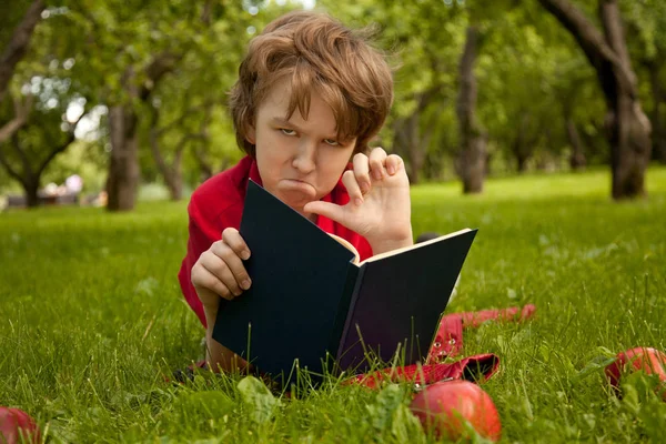 Triste aburrido sorprendido adolescente chico leyendo un libro en el verde verano manzanos parque — Foto de Stock