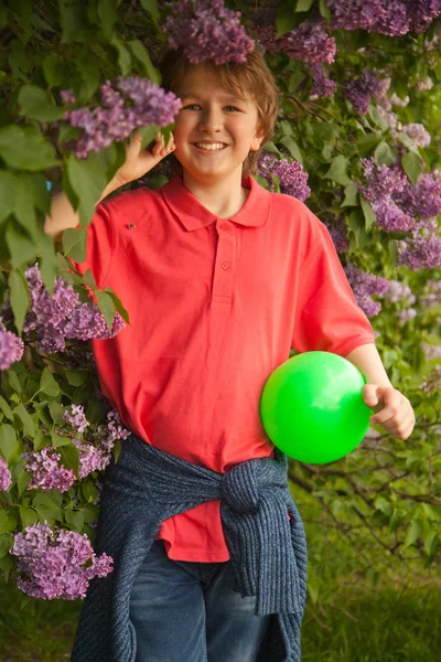Deportivo feliz niño sonriente con bola verde jugando en el parque de flores lila solo al aire libre —  Fotos de Stock