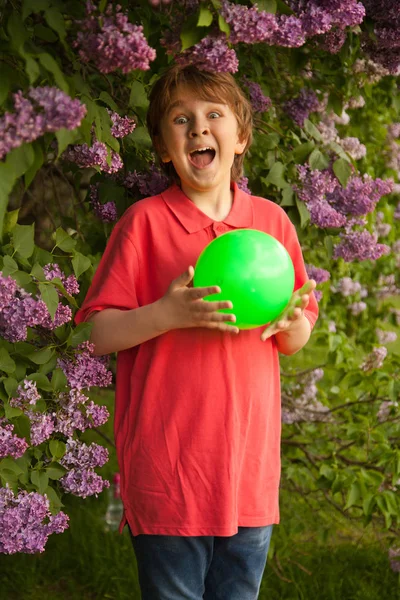 Deportivo feliz niño sonriente con bola verde jugando en el parque de flores lila solo al aire libre —  Fotos de Stock
