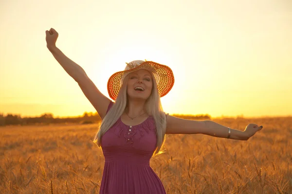 beautiful happy blonde girl with white straw hat standing in the wheat field in the sunset