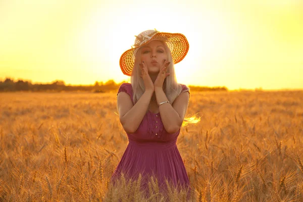 Mooie gelukkig blond meisje met witte stro hoed staan op het gebied van tarwe in de zonsondergang — Stockfoto