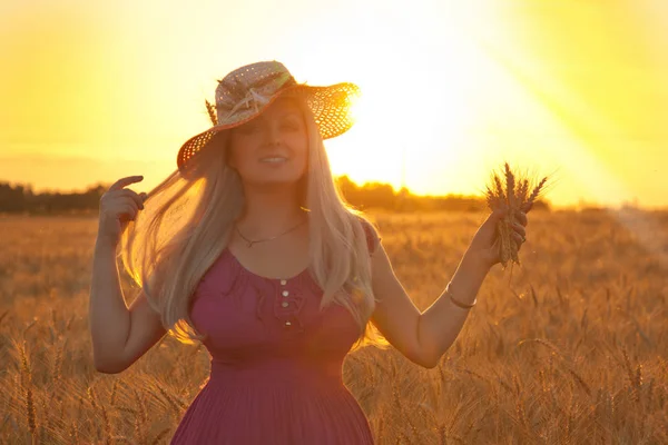 Mooie gelukkig blond meisje met witte stro hoed staan op het gebied van tarwe in de zonsondergang — Stockfoto