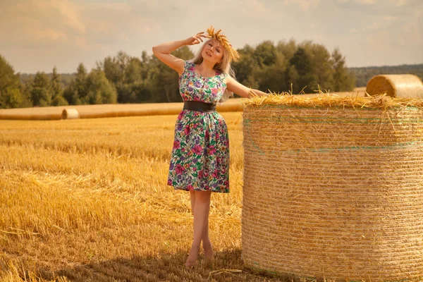 Prachtige landelijke vrouw bloem jurk en tarwe kroon staande in de buurt van droge ronde stro hooiberg dragen in het gele veld — Stockfoto