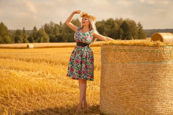 Prachtige landelijke vrouw bloem jurk en tarwe kroon staande in de buurt van droge ronde stro hooiberg dragen in het gele veld — Stockfoto