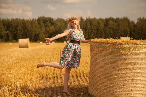 Belle femme rurale portant robe de fleur et couronne de blé debout près de paille ronde sèche meule de foin dans le champ jaune — Photo