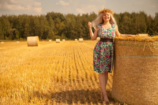 Krásné venkovské ženy nosí květinové šaty a pšenice koruny stojí poblíž suché kulaté slámy sena v žlutém poli — Stock fotografie