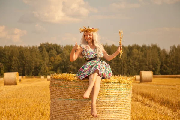 Beautiful rural woman wearing flower dress and wheat crown standing near dry round straw haystack in the yellow field — Stock Photo, Image