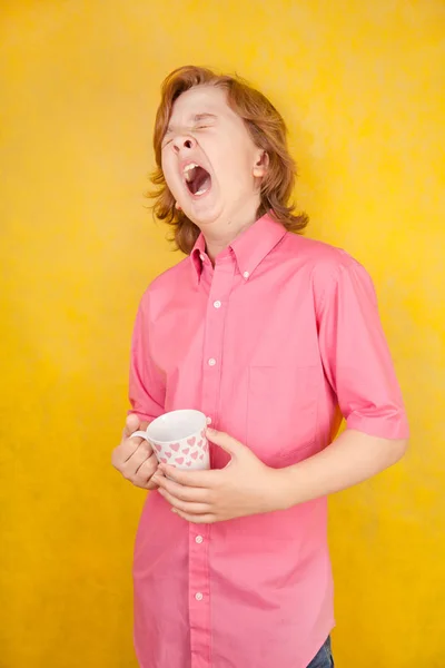 Tiener dragen roze shirt staande op de gele achtergrond in de studio en geeuwen met kopje koffie in de ochtend — Stockfoto