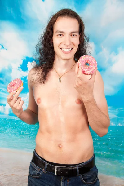 Hete sexy jonge lange haren man roze smakelijke donut eten op de blauwe studio achtergrond — Stockfoto