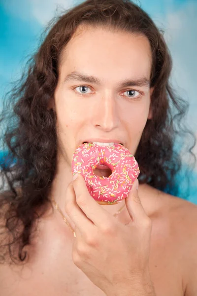 Hete sexy jonge lange haren man roze smakelijke donut eten op de blauwe studio achtergrond — Stockfoto