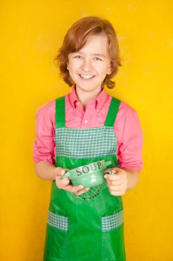 çorba tabağı ve bir kaşık ile yeşil mutfak önlük ayakta giyen güzel çocuk