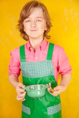 çorba tabağı ve bir kaşık ile yeşil mutfak önlük ayakta giyen güzel çocuk