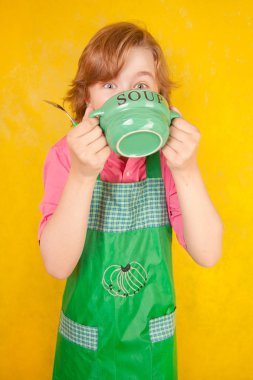 çorba tabağı ve bir kaşık ile yeşil mutfak önlük ayakta giyen güzel çocuk