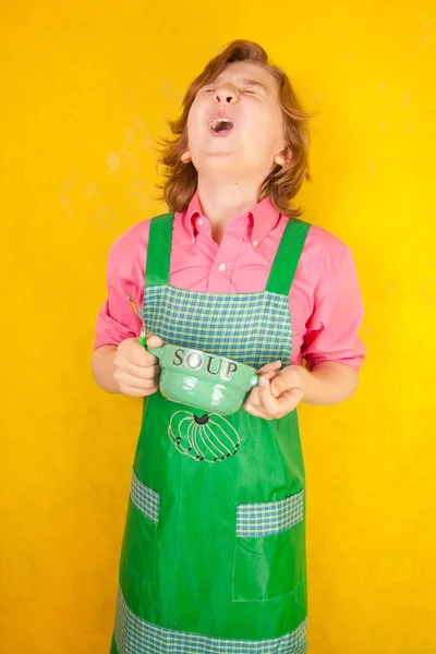 Ωραίο παιδί φοράει πράσινη κουζίνα ποδιά στέκεται με βαθύ πιάτο και ένα κουτάλι — Φωτογραφία Αρχείου