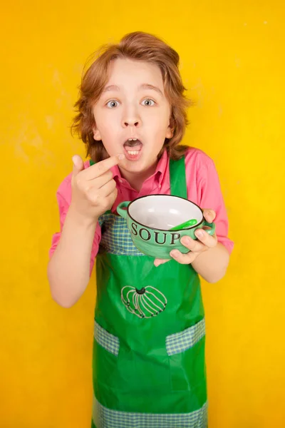 Ωραίο παιδί φοράει πράσινη κουζίνα ποδιά στέκεται με βαθύ πιάτο και ένα κουτάλι — Φωτογραφία Αρχείου