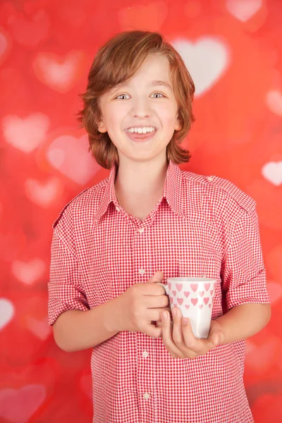 Χαριτωμένο ευτυχισμένη χαμογελαστό αγόρι με λευκό φλιτζάνι σε κόκκινη καρδιά studio φόντο μόνος — Φωτογραφία Αρχείου