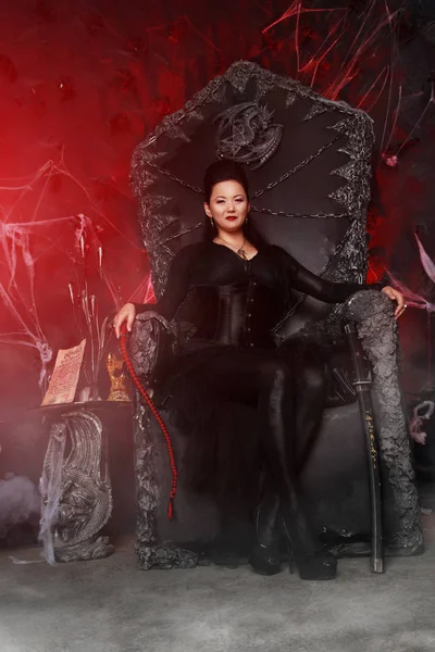 Charming dark evil witch woman wearing stylish gothic dress and sitting on the huge black queen chair — Stock Photo, Image