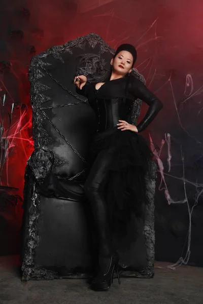 Encantadora mulher bruxa do mal escuro vestindo vestido gótico elegante e sentado na enorme cadeira rainha preta — Fotografia de Stock