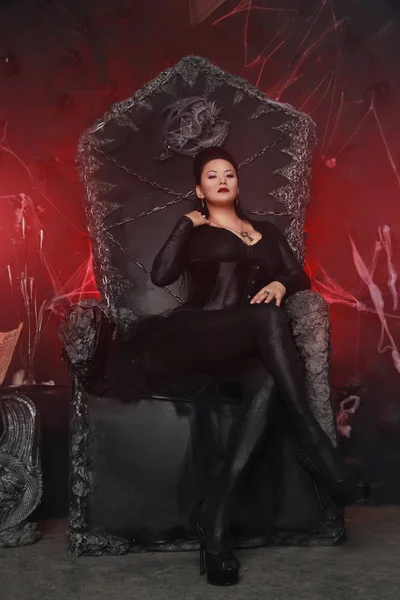 Charming dark evil witch woman wearing stylish gothic dress and sitting on the huge black queen chair — Stock Photo, Image