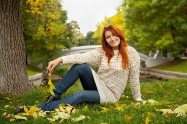 sıcak beyaz kazak giyen ve ağaç parkta yere yakın oturan güzel sonbahar Kızıl saçlı kadın