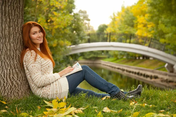 Bonita outono ruiva mulher vestindo camisola branca quente e sentado perto da árvore no chão no parque — Fotografia de Stock