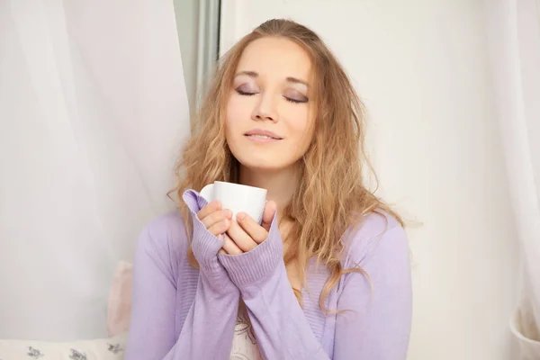 Pretty cute young woman wearing romantic purple sweater and long pink skirt and sitting on the white window sill — Stock Photo, Image