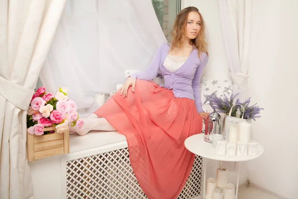 Bonita linda mujer joven con suéter púrpura romántico y falda larga rosa y sentado en el alféizar de la ventana blanca — Foto de Stock