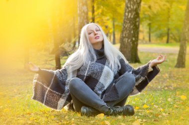 sıcak yün panço ve meditasyon rahatlama poz sonbahar parkta tek başına oturan kot giyen güzel sarışın kadın