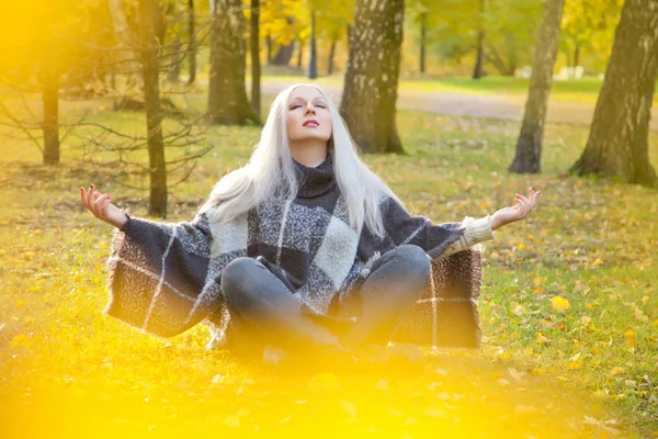 Vacker blond kvinna klädd i varm ull poncho och jeans som sitter i meditation avkoppling Posen i parken höst ensam — Stockfoto