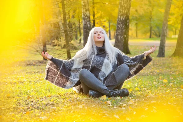 Vacker blond kvinna klädd i varm ull poncho och jeans som sitter i meditation avkoppling Posen i parken höst ensam — Stockfoto