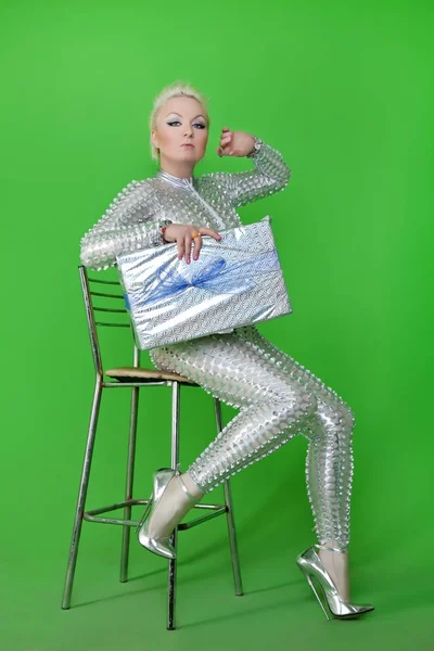 Estranho futurista loira mulher vestindo prata catsuit e sentado na cadeira com presente caixa de Natal no fundo cromado verde sozinho — Fotografia de Stock