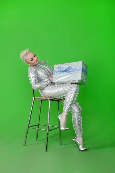 Strange futuristic blonde woman wearing silver catsuit and sitting on chair with gift christmas box on green chromakey background alone — Stock Photo, Image