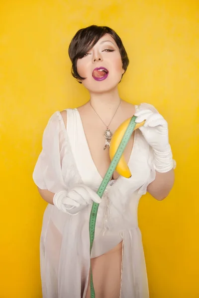 Pretty sexual brunette woman with sweet banana and measurement tape standing on the yellow studio background alone — Stock Photo, Image