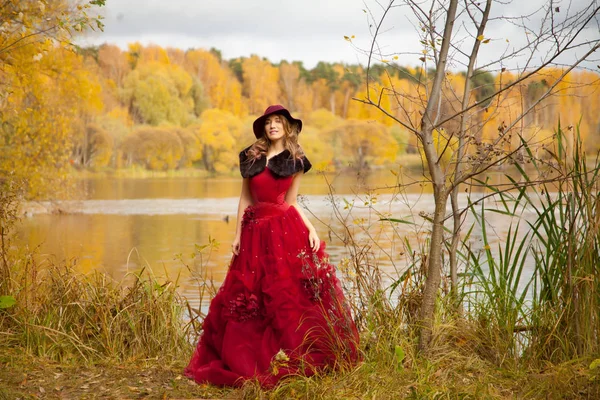 Piękna młoda kobieta w czerwonej sukience długie ciemne i w parku, jesień w pobliżu jeziora i żółty upadek drzew — Zdjęcie stockowe