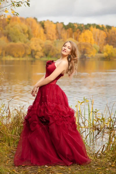 Bela jovem mulher vestindo longo vestido vermelho escuro e de pé no parque de outono perto do lago e árvores de queda amarelas — Fotografia de Stock