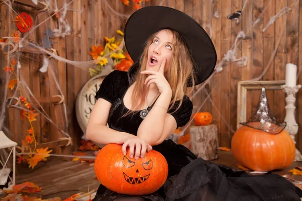 Hermosa mujer bruja clásica con vestido de terciopelo bruja negro y sombrero puntiagudo, con calabazas de halloween mal naranja y tela de araña — Foto de Stock